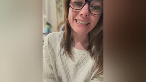 A video of a smiling Caucasian woman with glasses and long brown hair, wearing a white cable-knit sweater. The background is blurred, showing a bathroom with a green and blue toiletry bottle.