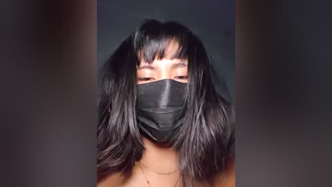 A video of an Asian woman with long, black hair, wearing a black face mask, and a plain black top. She has straight bangs and is gazing directly at the camera.