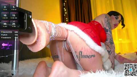 Video of a tattooed woman in a red Santa outfit, kneeling on a fluffy carpet, with a large dildo penetrating her anus, in a warmly-lit room with curtains.