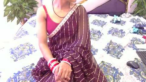 Video of a woman in a maroon saree with white stripes, wearing a red blouse, seated on a bed with a blue and white patterned bedspread. The room has green plants and scattered objects.