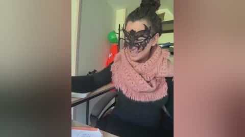 Video of a woman with dark hair in a bun, wearing a black lace masquerade mask, a pink crocheted scarf, and a black top, seated in a dimly lit room with a red Christmas ornament and a green balloon.