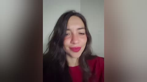 A video of a smiling woman with long, wavy dark hair, wearing red lipstick, a red top, and a black jacket, set against a blurred, neutral background.