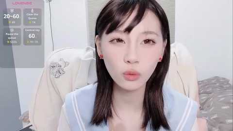 Media: Video of a young Asian woman with straight black hair and fair skin, wearing a blue sailor collar uniform. She sits on a bed with a cream-colored pillow and a grey blanket, in a simple, minimalistic room.
