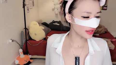 Video of a young woman in a white nurse uniform with a pink headband, wearing a white face mask and a black vibrator. Background shows a messy room with stuffed animals and a red bed.