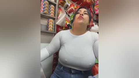 A video of a curvy Latina woman with medium brown skin and dark hair, wearing a tight, white ribbed top and high-waisted jeans, leaning back against a cluttered snack shelf in a dimly lit convenience store.