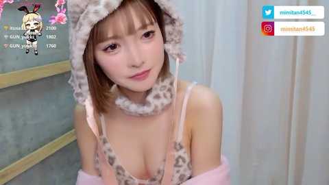 Video of a young Asian woman with fair skin and straight brown hair, wearing a leopard-print hooded top and matching scarf, indoors. Background features light curtains and a wall with a floral design.
