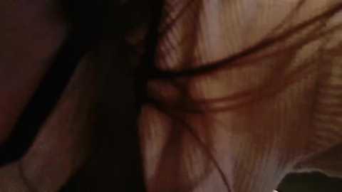 A close-up video shows a woman's face with long, wavy black hair partially covering her cheek and nose. She wears black makeup and has a fair skin tone. The image is dimly lit, creating a moody atmosphere.