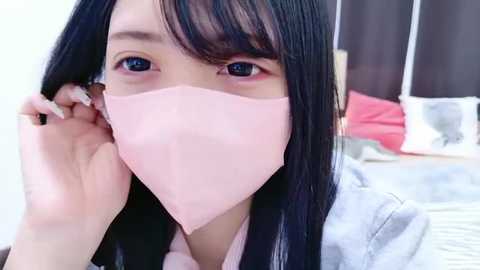 Video of an Asian woman with long black hair, wearing a pink face mask, adjusting it while sitting on a bed with red and white pillows.