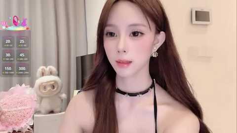 Video of a young Asian woman with long brown hair, wearing a black choker, earrings, and a black top. She's indoors, next to a gray curtain, a pink bunny plush, and a white wall.