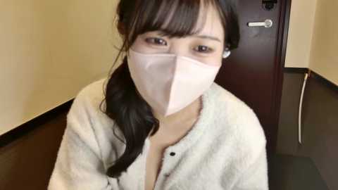 Video of an East Asian woman with long black hair, wearing a light pink surgical mask and a white bathrobe, standing indoors near a wooden door.