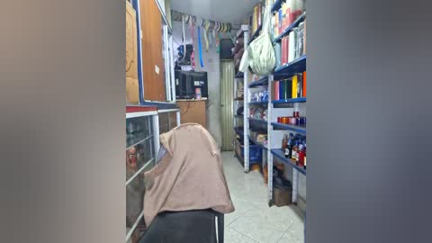 A video of a small, cluttered room with beige walls and blue shelves. A beige towel is draped over a chair. The room is dimly lit, with various personal items and household goods scattered about.