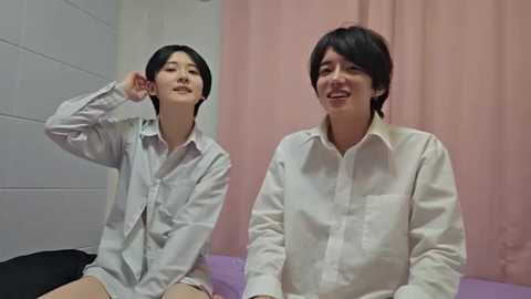 Video of two East Asian women in white button-up shirts, one with short black hair and another with long black hair, sitting side by side on a bed with a purple blanket, against a pink curtain background.