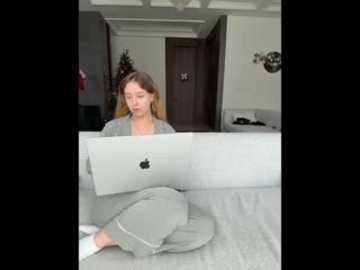 Video of a blonde woman in a gray cardigan and pajama pants, sitting on a white couch, working on an open MacBook, with a gray hat beside her. Background shows a modern living room with white walls and Christmas decorations.