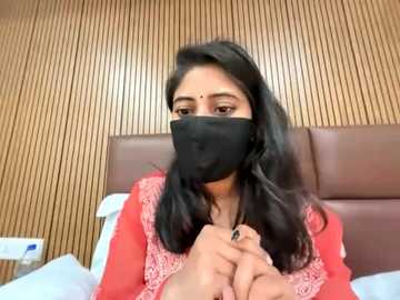 A video of a young woman with long black hair, wearing a red saree and black face mask, sitting on a bed with a wooden headboard.