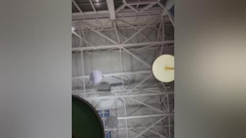 A video showing an indoor basketball court with a high, white, metallic ceiling and a large, circular, yellow light fixture. The court's green surface and a white net are partially visible.