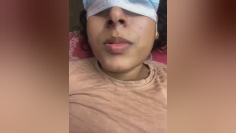 Video of a woman with medium brown skin, wearing a beige t-shirt and blue face mask, lying on a red and pink floral-patterned pillow.