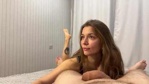 Video of a nude Caucasian woman with long brown hair, medium build, lying on a bed with white sheets and white walls. She is smiling, with her legs raised and crossed, showcasing her tattooed arm.