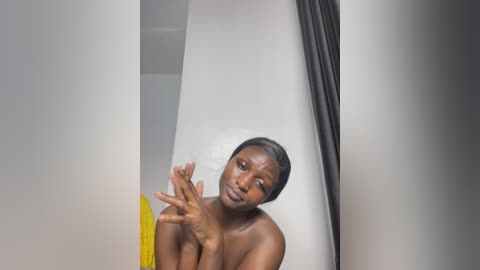 A video of a topless Black woman with a slender physique, medium-length black hair, and a serene expression, sitting in a modern, minimalist room with white walls and a yellow towel.