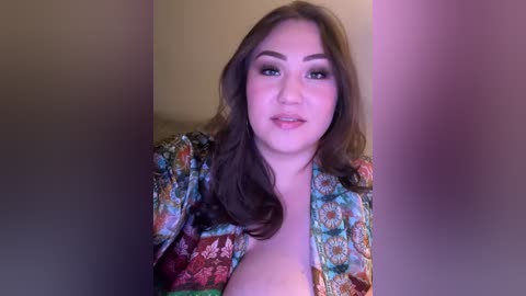 Media: Video of a young Asian woman with long, wavy brown hair, wearing a colorful, floral-patterned robe, standing in a blurred, beige-walled room. She has a soft, round face with a slight smile.