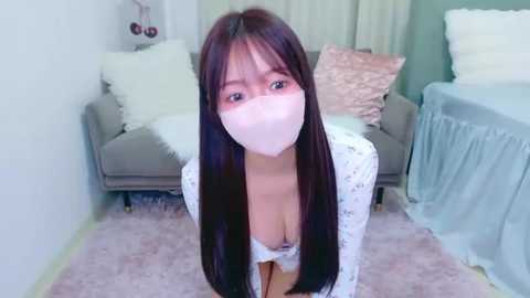 Video of an Asian woman with long black hair, wearing a white mask and a low-cut floral-patterned top, kneeling on a pink shag rug in a pastel bedroom.