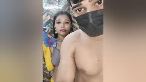 Video of an Indian woman with fair skin, wearing a yellow sari, blue blouse, and a face mask, standing beside a shirtless man with a black mask. Background includes a colorful, patterned cloth.
