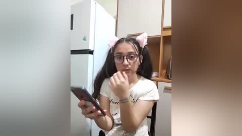 A young girl with long dark hair, wearing glasses, white t-shirt, and cat ears, sits on a wooden chair, holding a smartphone, in a kitchen with a white fridge and cabinets.