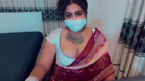 Video of a curvy, light-skinned woman with dark hair in a bun, wearing a white bra, red sari, and silver necklace, seated on a bed with beige and black curtains in the background.