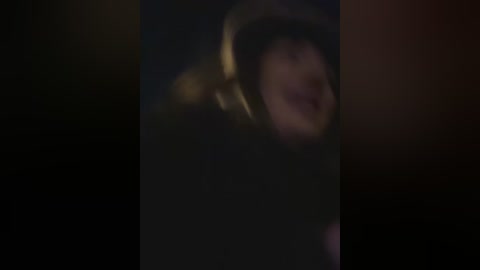 A dimly lit, blurred video of a person wearing a black hoodie with headphones on, their face obscured, against a dark, indistinct background.