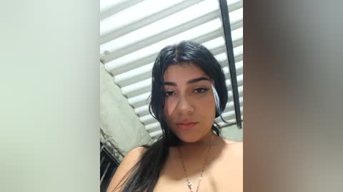 Video of a young woman with long black hair, light brown skin, and dark eyes, standing in a dimly lit room with white blinds and a beige wall. She is topless, revealing her bare shoulders and chest, wearing a delicate necklace.