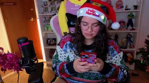 Video of a young woman in a festive Santa hat, wearing a colorful sweater, playing with a smartphone. Background shows a cozy room with shelves and a decorated Christmas tree.