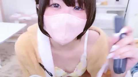 A video of a young, fair-skinned Asian woman with short, dark brown hair, wearing a yellow robe and a white face mask, holding a blue remote control, in a bright, modern room.