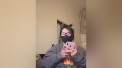 A young Asian woman with a chubby face and short dark hair takes a selfie in a dimly lit room. She wears a black mask, glasses, and a gray sweatshirt with a colorful design.