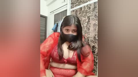 A video of a young woman with long, dark hair, wearing a red lace bra, black mask, and sheer red robe, seated in a bathroom with marble walls.