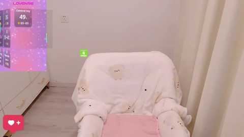 A video of a cozy, pastel-themed room featuring a white armchair adorned with teddy bear cushions, with a soft, plush blanket draped over it. The background includes a white bed and a green digital screen displaying a game interface.