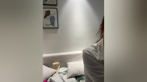 Video of a cozy, dimly lit room with a white bed, beige pillows, and abstract art on the wall. A person with long brown hair is partially visible.