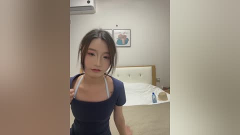 Video of an East Asian woman with fair skin, wearing a navy blue dress, standing in a minimalist bedroom with a white bed, framed artwork, and an air conditioner.