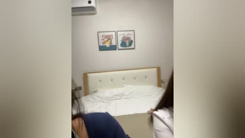 Video of a small, dimly lit room with a white bed, two framed posters on the wall, and a person in a blue shirt leaning over the bed.