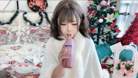 Video of a young Asian woman with shoulder-length brown hair, wearing a white robe, drinking from a pink bottle labeled \"Aqua.\" She's in a festive room with a decorated Christmas tree and holiday-themed decor.