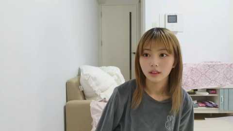 Video of a young East Asian woman with straight, light brown hair, wearing a gray T-shirt, sitting in a beige living room with a white door and a pink quilted pillow.