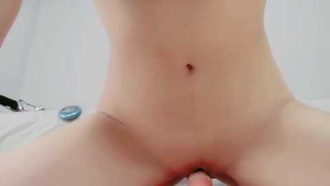 Video of a nude, light-skinned person with visible genitals, sitting with legs spread, showing a small scar on the inner thigh. Background features a white wall and a blue object in the lower left corner.