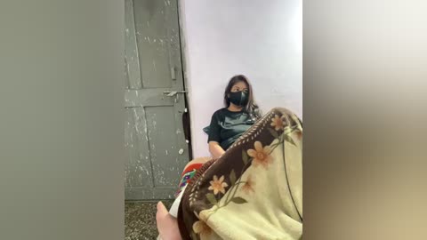 Media: Video of a woman with shoulder-length hair, wearing a black mask and dark t-shirt, sitting in a dimly lit room with a door and peeling paint.