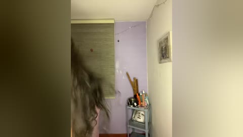 A video captures a blurry, partially obstructed figure in a dimly lit room. The room has light lavender walls, a window with a closed blind, a cluttered desk, and a framed picture. The atmosphere is somewhat eerie and mysterious.