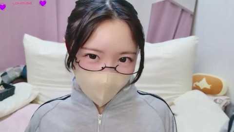 Video of an East Asian woman with glasses, wearing a beige mask, gray jacket, and dark hair, lying on a bed with white pillows and pastel-colored bedding in a cozy room.