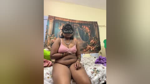Media: Video of a plus-sized Black woman with dark skin, wearing a pink bra and panties, sitting on a bed with floral sheets, surrounded by a colorful tapestry.