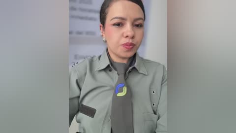 Video of a young woman with short dark hair, wearing a gray shirt and tie, standing in a white office hallway with blurred text on the wall.