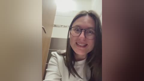 Video of a smiling woman with glasses and long brown hair, standing in a bathroom doorway, wearing a white shirt.