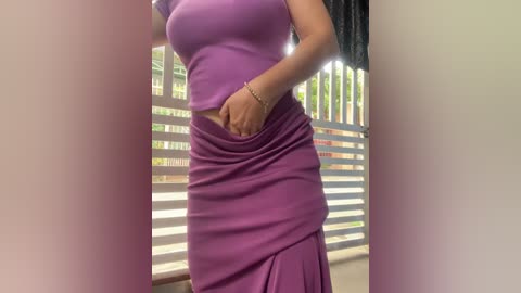 Video of a woman with medium-dark skin, wearing a tight, sleeveless, purple crop top and matching high-waisted, ruched skirt. She stands in a bright room with white walls and vertical blinds.