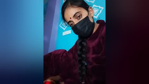 Video of a woman with dark skin, wearing a black mask, red dot on forehead, and long braided hair, in a teal room with white geometric patterns.
