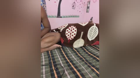 Video of a person sitting on a bed with a plaid blanket, wearing a blue-patterned top and shorts, in a pink bedroom with wall art and a leafy plant.