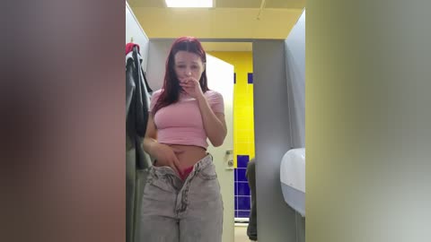 A video of a young woman with red hair, wearing a pink crop top and unbuttoned jeans, taking a selfie in a brightly lit bathroom with yellow and blue tiled walls.